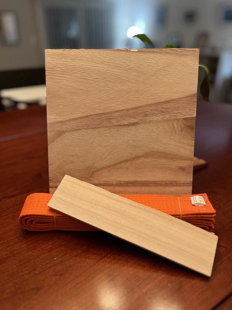 A broken board sits on top of a table. The larger part of the board is standing up (propped up). In front of it is a folded, orange taekwondo belt. Leaning on the belt is the much smaller part of the broken board.